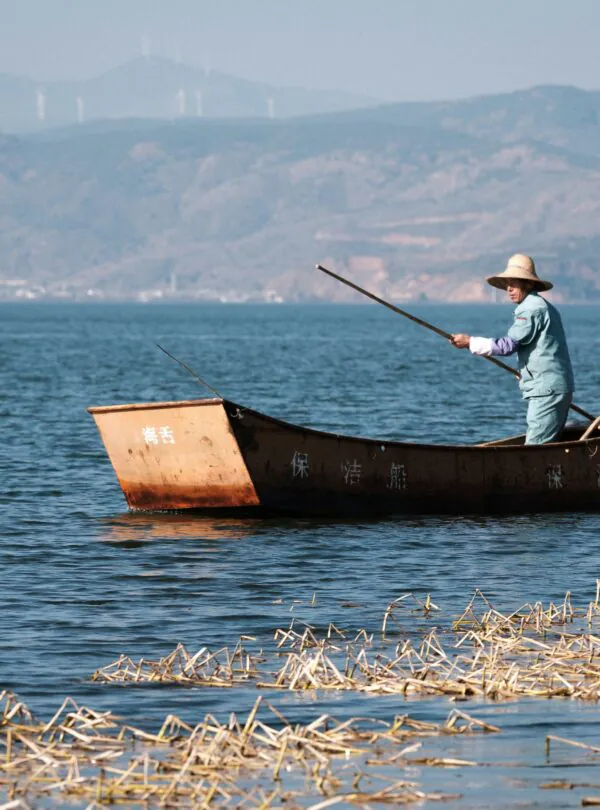 8-Day Yunnan Culinary Cultural Adventure, china, china tour, Person rowing a small rust-colored boat on a calm blue lake, wearing a wide-brimmed straw hat and light clothing, with distant mountains in the background