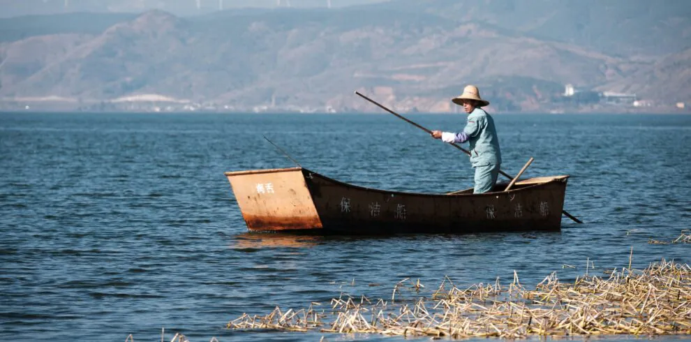 8-Day Yunnan Culinary Cultural Adventure, china, china tour, Person rowing a small rust-colored boat on a calm blue lake, wearing a wide-brimmed straw hat and light clothing, with distant mountains in the background