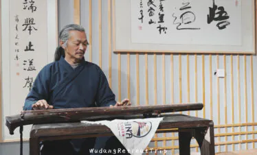 wudang taichi retreat, taoist, hubei, china, chinaexplorertour, Elderly man with gray hair in a dark blue traditional robe playing a Chinese string instrument (guzheng) at a wooden table, indoors with calligraphy panels on the wall
