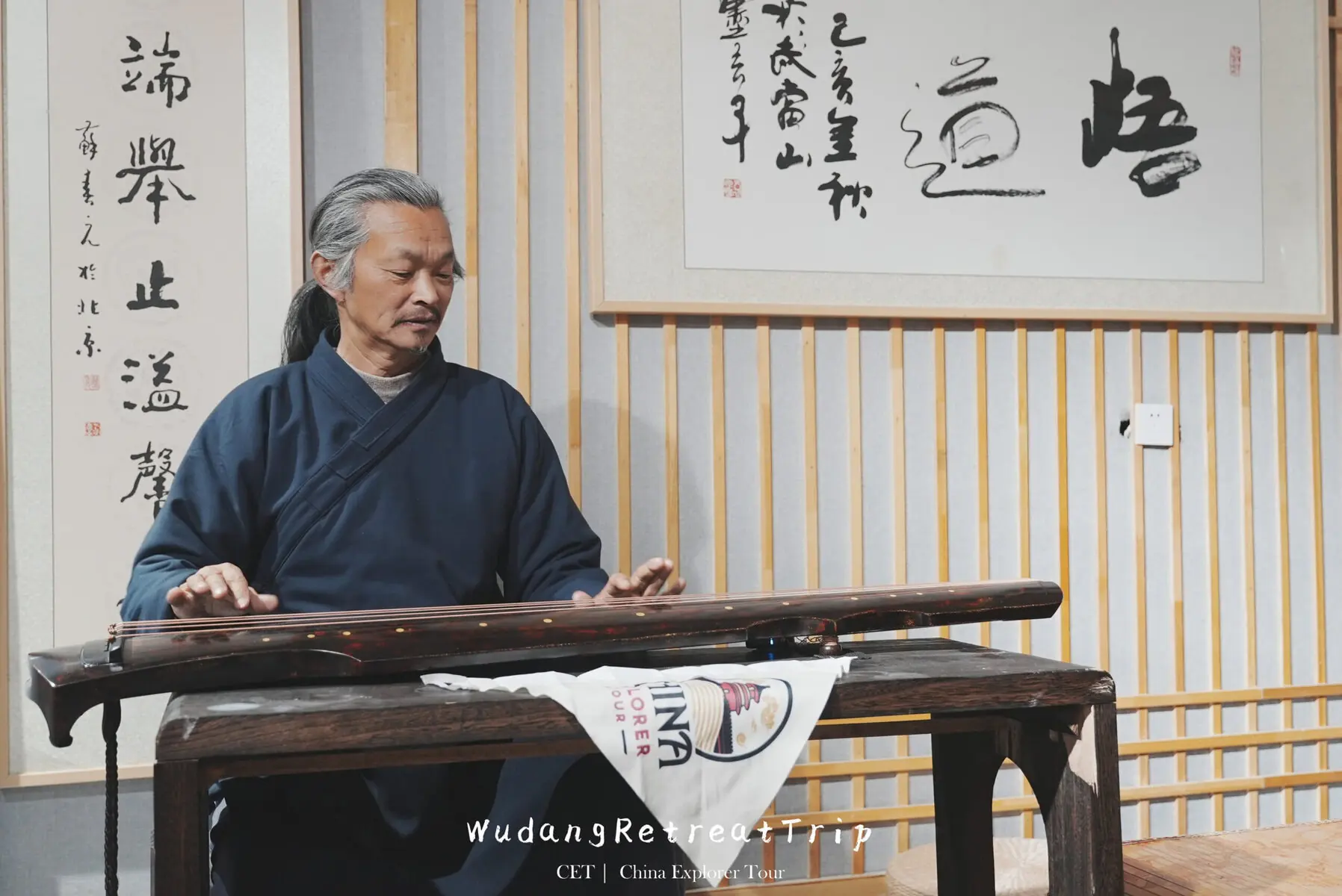 wudang taichi retreat, taoist, hubei, china, chinaexplorertour, Elderly man with gray hair in a dark blue traditional robe playing a Chinese string instrument (guzheng) at a wooden table, indoors with calligraphy panels on the wall