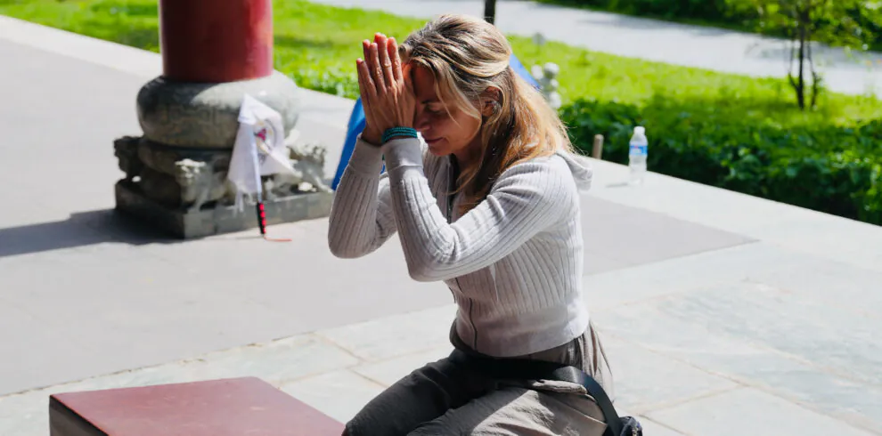 wutai retreat, shanxi, chinatrip, Woman kneeling in a peaceful outdoor setting, hands pressed together in prayer, eyes closed, with a retreat banner visible at the bottom.
