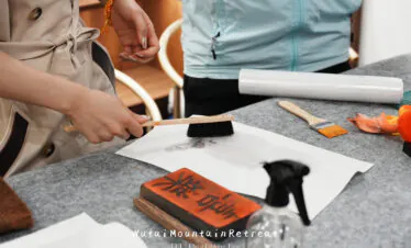 wutai retreat, chinatrip, Hands painting on white paper with a brush, ink block and rolled paper on a gray table; orange bracelet visible on wrist.