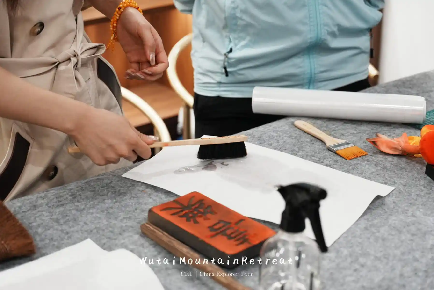 wutai retreat, chinatrip, Hands painting on white paper with a brush, ink block and rolled paper on a gray table; orange bracelet visible on wrist.