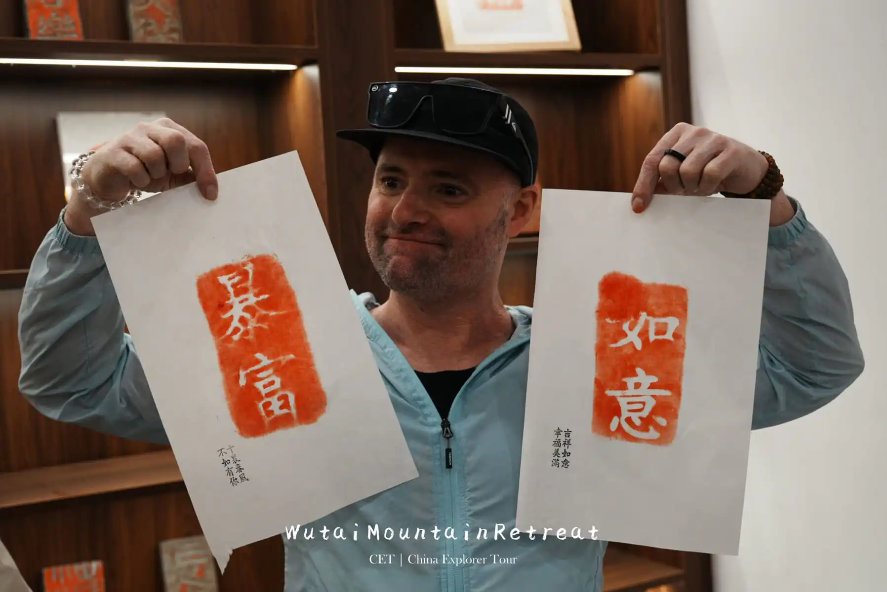 wutai retreat, shanxi, chinatrip, Smiling man in a light blue jacket holds up two calligraphy prints with orange-red seals and white Chinese characters.