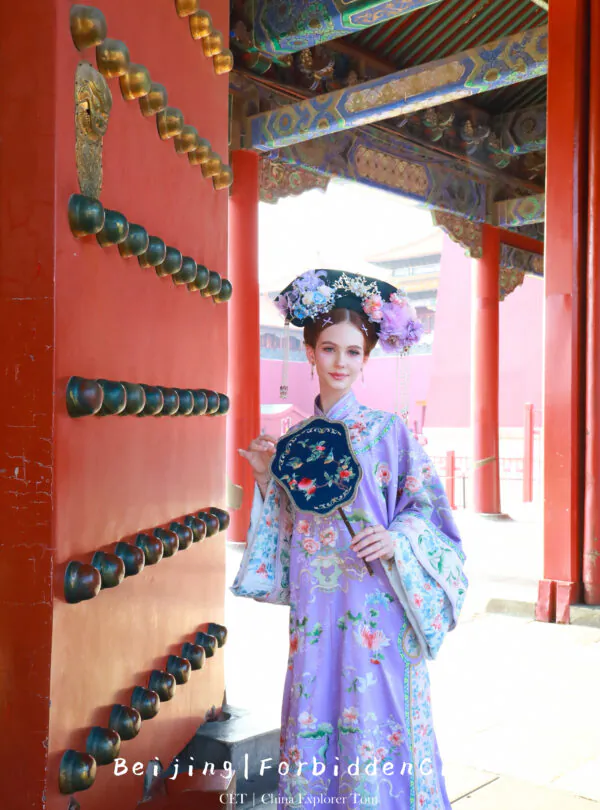 hanfu photoshoot, the forbidden city, beijing, qing dynasty, female, solo, chinatravel, Woman in a lavender traditional Chinese dress with a floral headdress posing beside a red ornate gate.