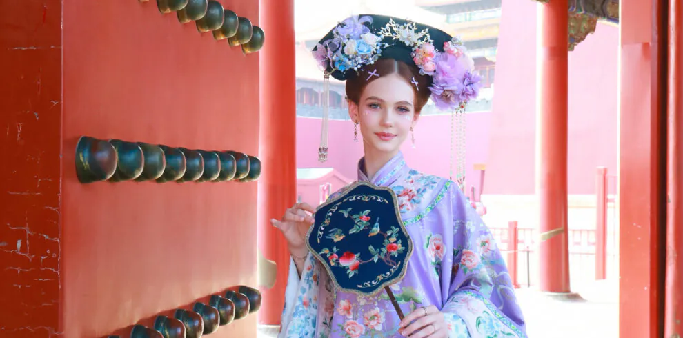 hanfu photoshoot, the forbidden city, beijing, qing dynasty, female, solo, chinatravel, Woman in a lavender traditional Chinese dress with a floral headdress posing beside a red ornate gate.
