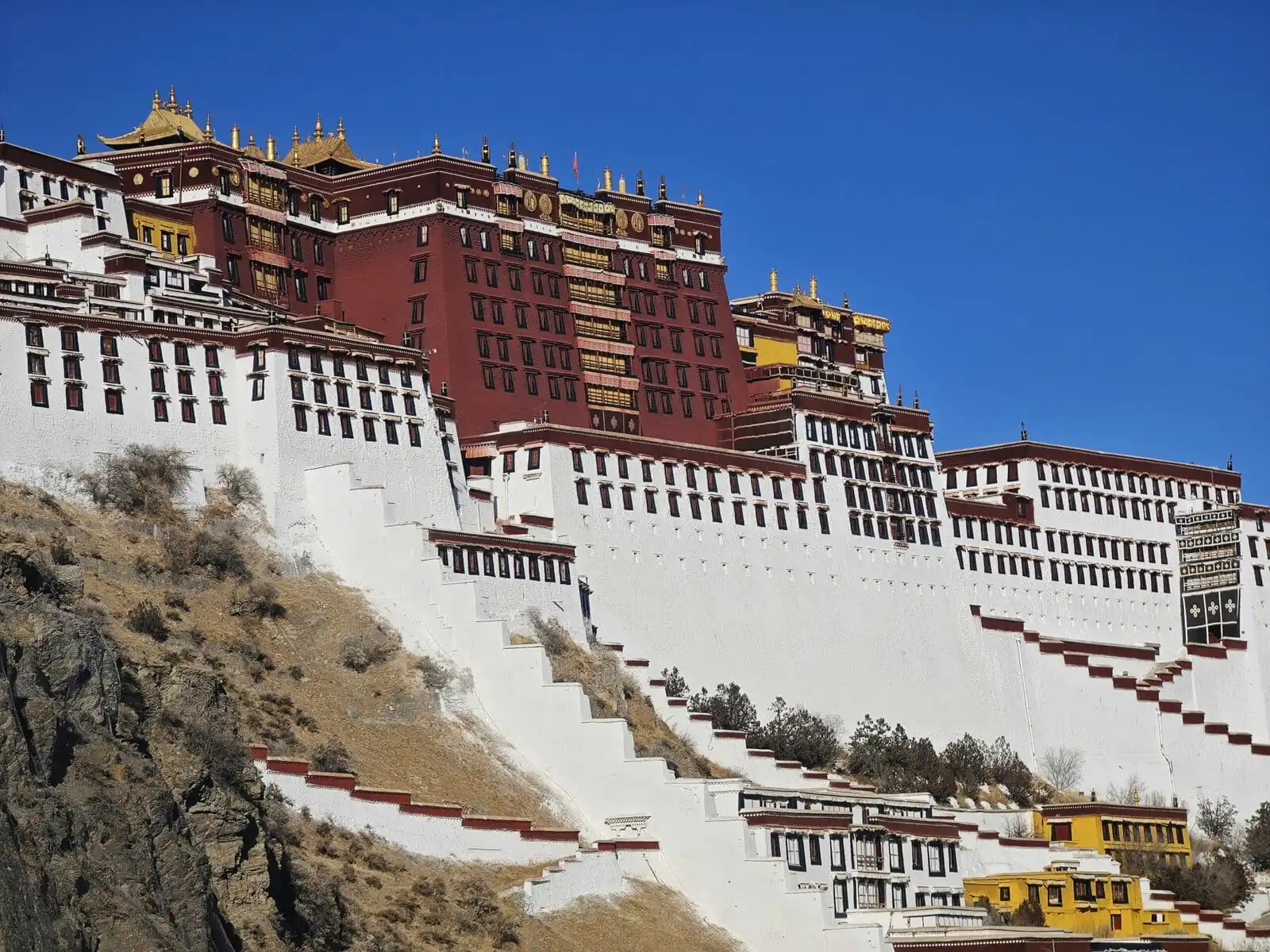 Potala Palace Tibet, china, china tour, Potala Palace in Lhasa perched on a hillside with white walls, red and gold buildings, and a clear blue sky above.