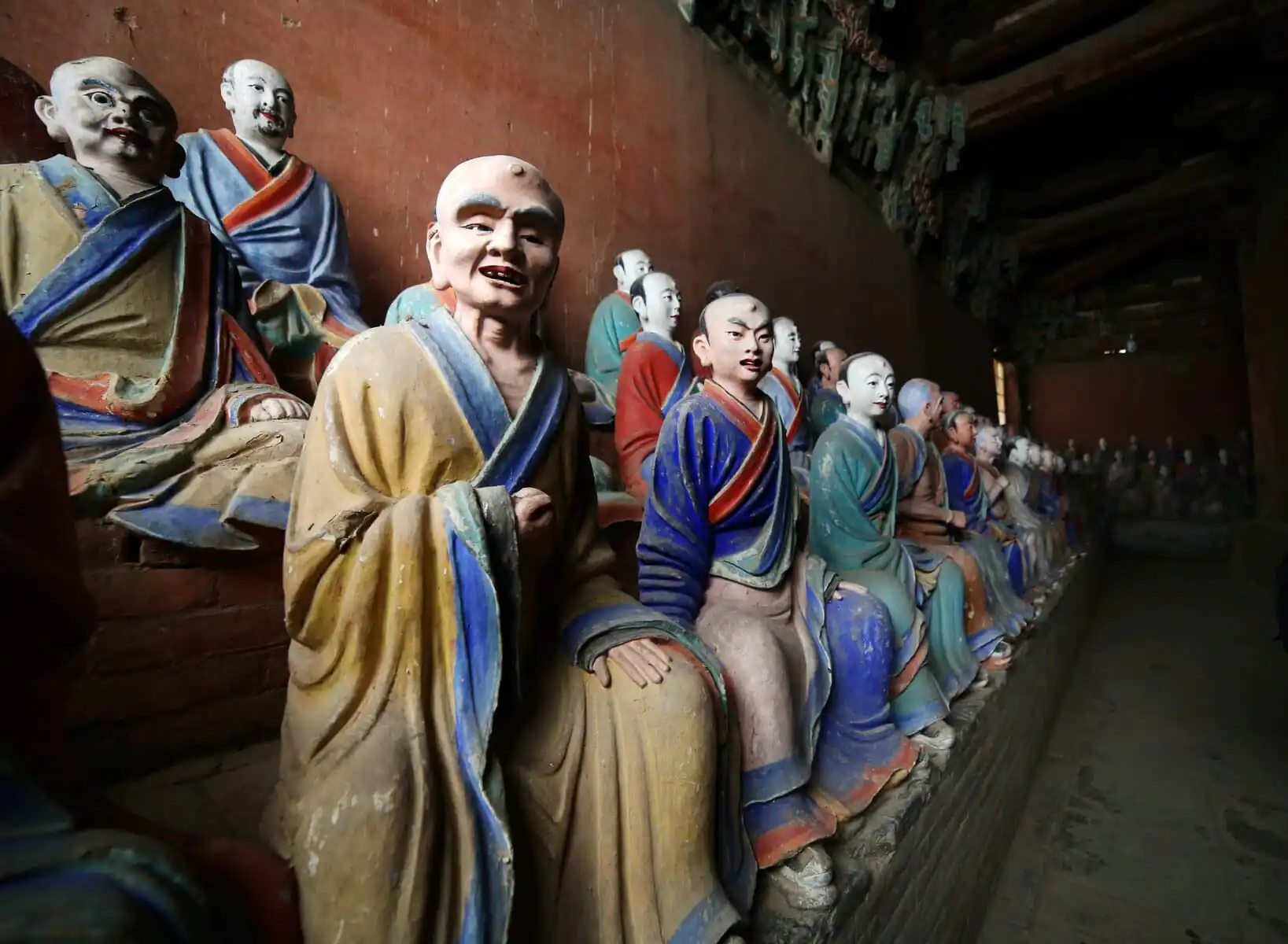 Foguang Temple, Mount Wutai, shanxi, china, china tour, Line of colorful seated Buddhist statues along a wooden wall inside a dim temple, facing forward with robes in yellow, blue, and green.