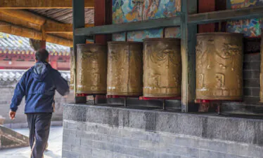 Tayuan Temple – Chan Hall, wutai retreat, shanxi, china, china tour, Person in a blue jacket walking past a row of large bronze prayer wheels inside a temple, with colorful murals overhead