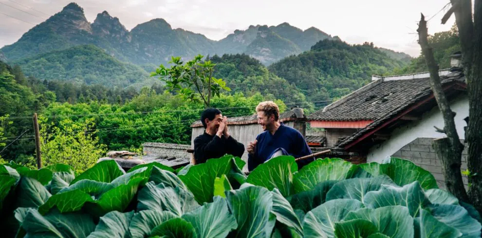 5-Day Wudang Mountain Tai Chi Retreat, hubei, china tour, Two men chat in a rural vegetable garden with oversized leafy greens in the foreground and mountains and traditional houses in the background.