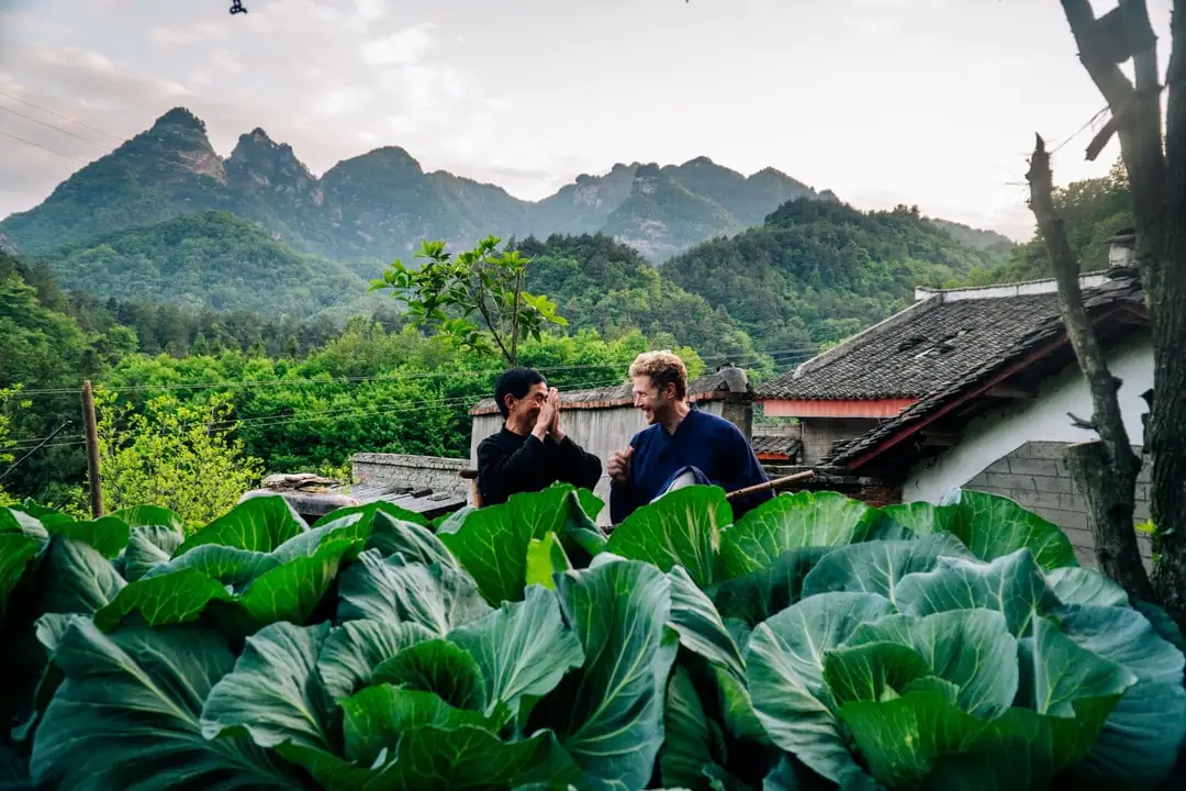 5-Day Wudang Mountain Tai Chi Retreat, hubei, china tour, Two men chat in a rural vegetable garden with oversized leafy greens in the foreground and mountains and traditional houses in the background.