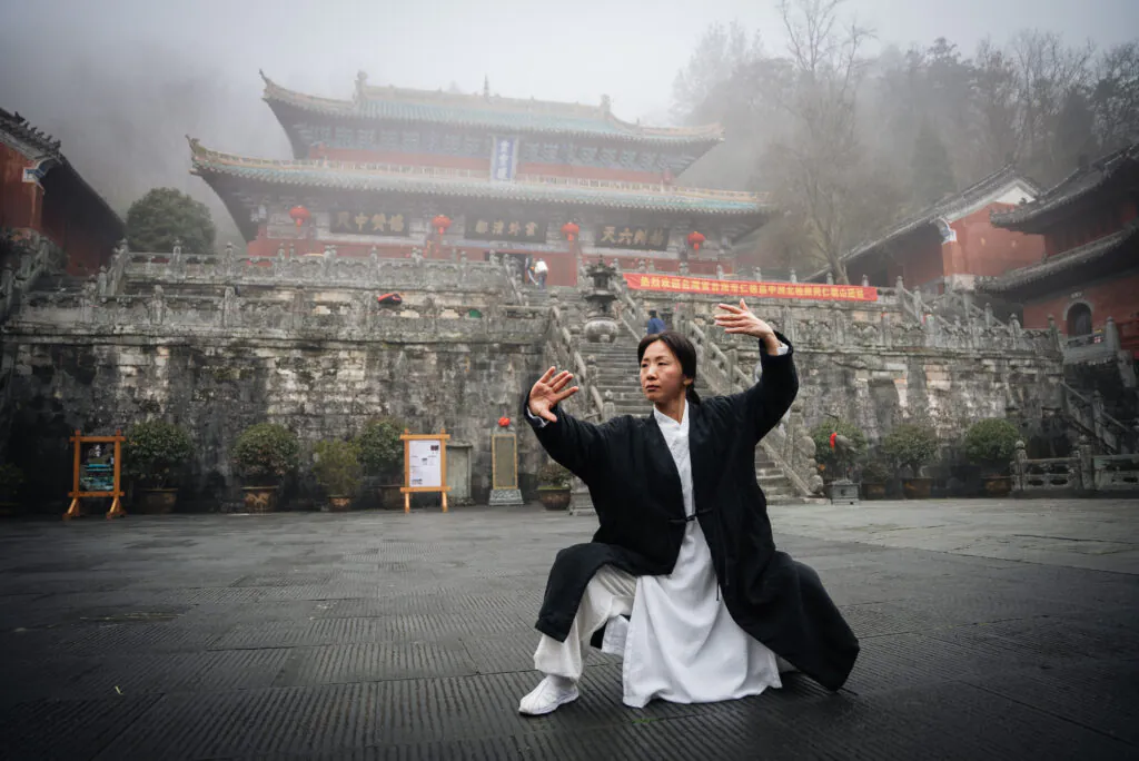 Wudang Mountain Tai Chi Retreat, hubei, china tour, Woman in white robe and black coat performs a martial-arts pose in a misty temple courtyard with stone steps behind.