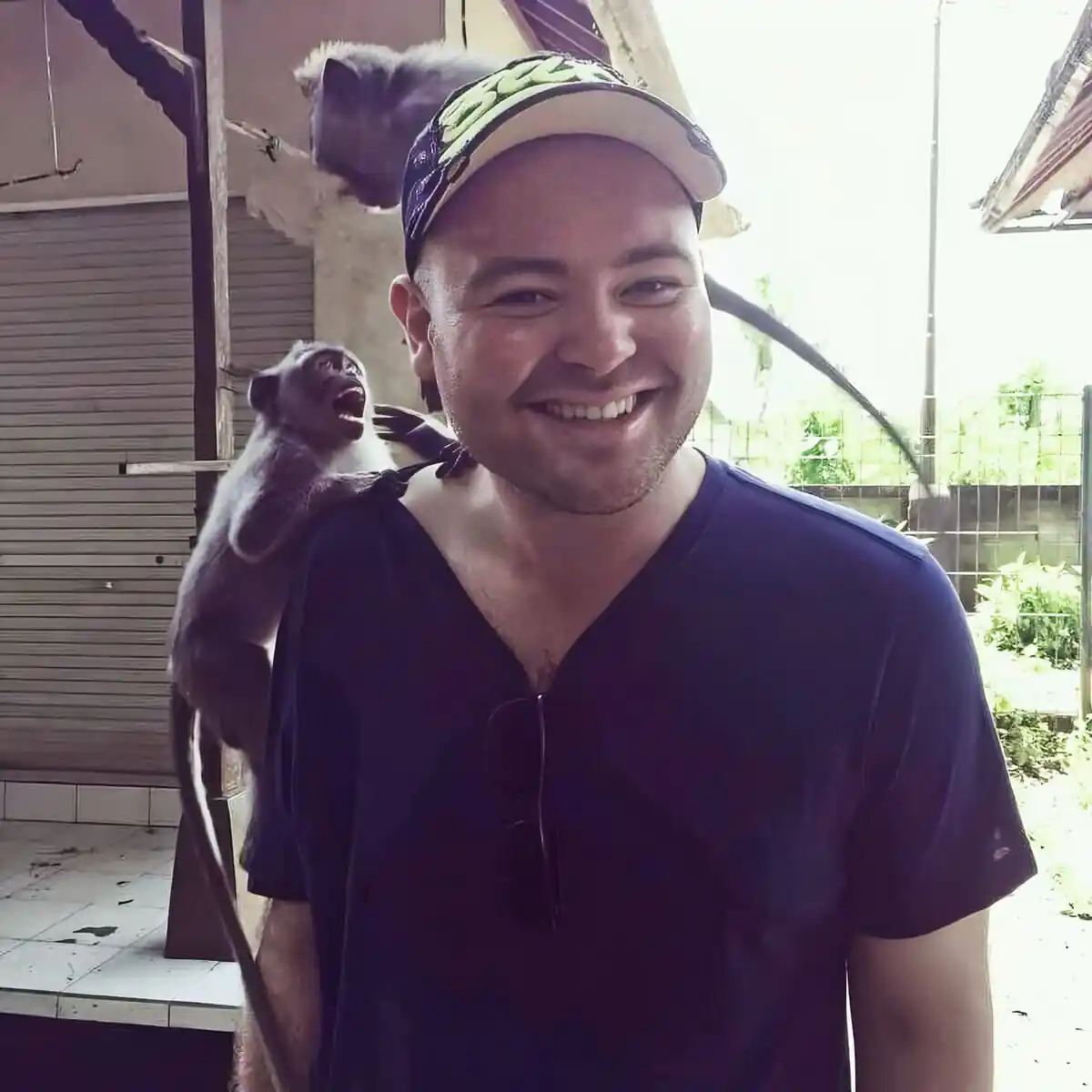 Smiling man in a navy T-shirt wearing a cap with sunglasses hanging from his shirt, a small monkey perched on his shoulder outdoors.