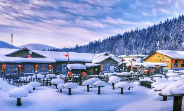 Enchanting Winter Wonderland: 3-Day Journey Through Harbin: China's Snow Kingdom, chinatrip, Snow-covered village with wooden houses and bright red lanterns along a snowy path. Nearby pine trees and hills in the background winter scene.