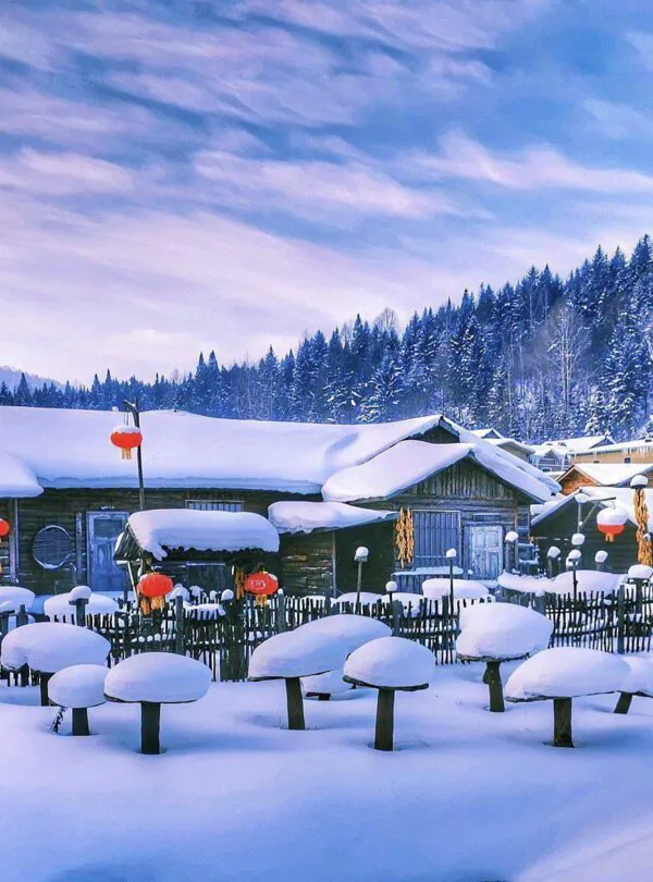 Enchanting Winter Wonderland: 3-Day Journey Through Harbin: China's Snow Kingdom, chinatrip, Snow-covered village with wooden houses and bright red lanterns along a snowy path. Nearby pine trees and hills in the background winter scene.