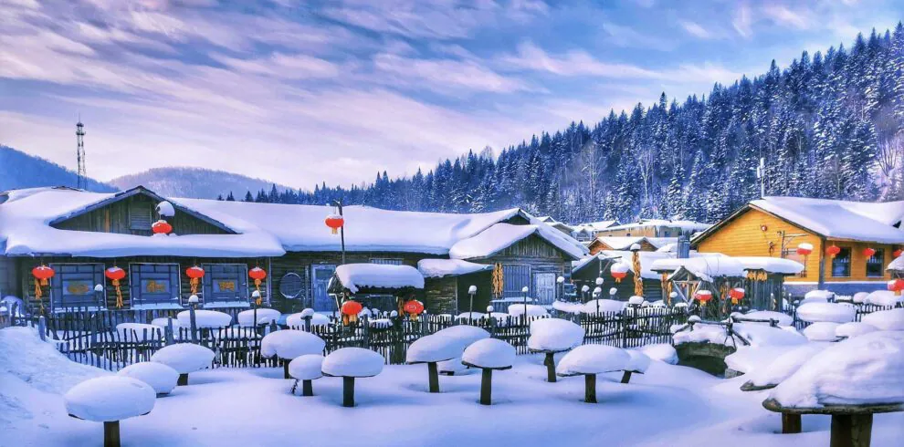 Enchanting Winter Wonderland: 3-Day Journey Through Harbin: China's Snow Kingdom, chinatrip, Snow-covered village with wooden houses and bright red lanterns along a snowy path. Nearby pine trees and hills in the background winter scene.