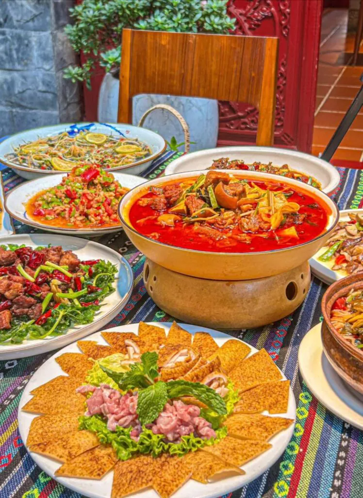 Yunnan Delicacy, china, china tour, Large red hot pot in the center with meat and vegetables, surrounded by multiple colorful dishes on a patterned tablecloth outdoors.