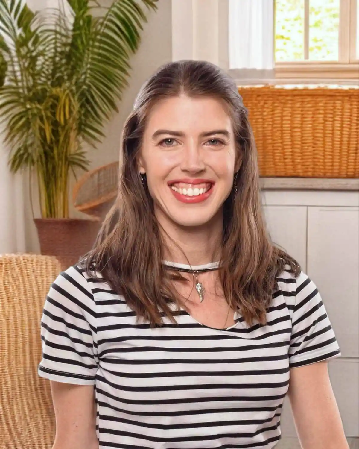 Smiling woman with brown hair in a black-and-white striped shirt, indoors with wicker furniture and a plant behind her.