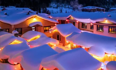 Enchanting Winter Wonderland: 3-Day Journey Through Harbin: China's Snow Kingdom, chinatrip, Snowy mountain village at night with warm orange lights glowing from windows and heavy snow on roofs under a starry blue sky.