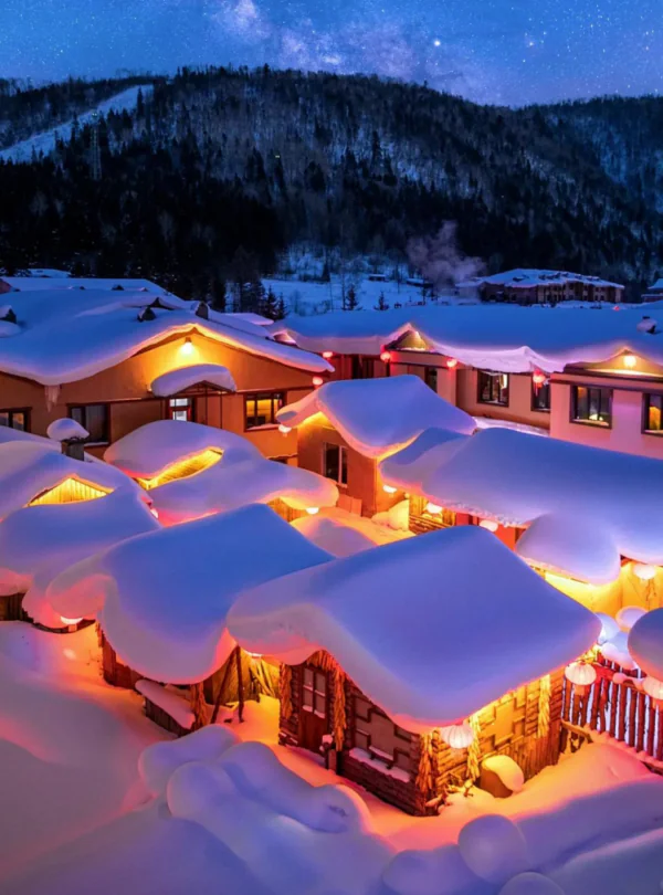 Enchanting Winter Wonderland: 3-Day Journey Through Harbin: China's Snow Kingdom, chinatrip, Snowy mountain village at night with warm orange lights glowing from windows and heavy snow on roofs under a starry blue sky.