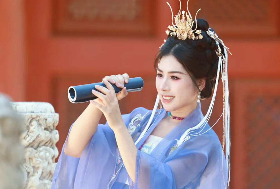 hanfu photoshoot, the forbidden city, beijing, female, solo, chinatrip, A woman wearing a blue traditional Chinese hanfu with an ornate gold headdress holding a rolled blue scroll like a microphone, speaking at a Beijing location.