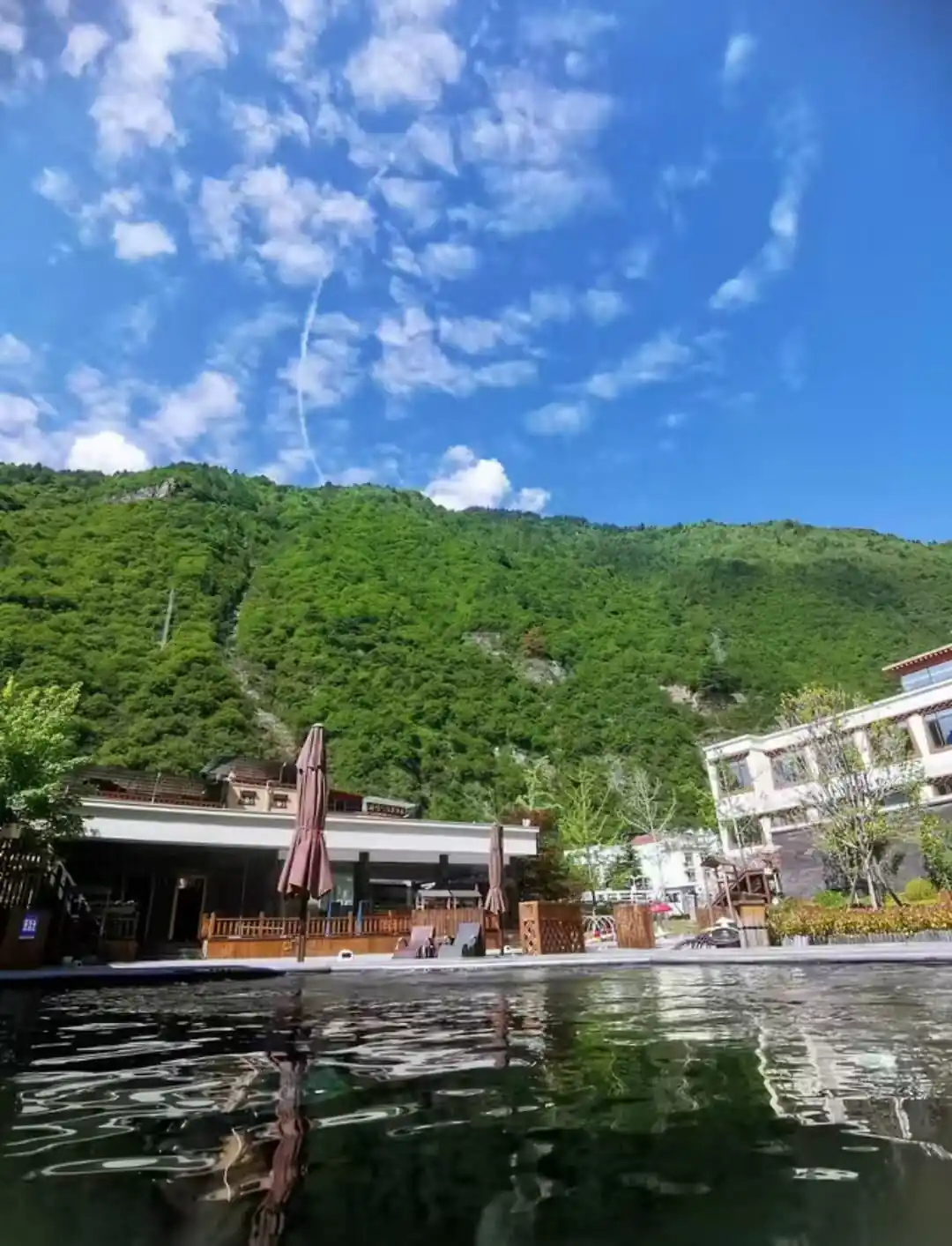Hot Spring, Bipenggou, 3-Days Explore Western Sichuan in Winter- Siguniang Mountain, Bipenggou, and Dagu Glacier, china tour, Lakeside resort with a wooden deck, brown umbrellas, and lounge chairs by calm water, set against green mountains under a blue sky with scattered clouds