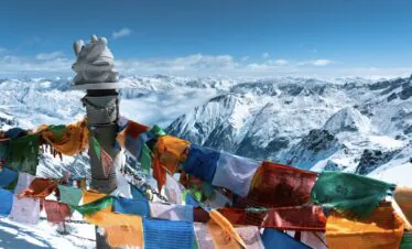 Bipenggou, 3-Days Explore Western Sichuan in Winter- Siguniang Mountain, Bipenggou, and Dagu Glacier, china tour, Colorful prayer flags flutter around a stone cairn atop a snowy mountain, with a vast range of snowcapped peaks in the background.