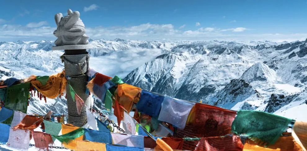 Bipenggou, 3-Days Explore Western Sichuan in Winter- Siguniang Mountain, Bipenggou, and Dagu Glacier, china tour, Colorful prayer flags flutter around a stone cairn atop a snowy mountain, with a vast range of snowcapped peaks in the background.