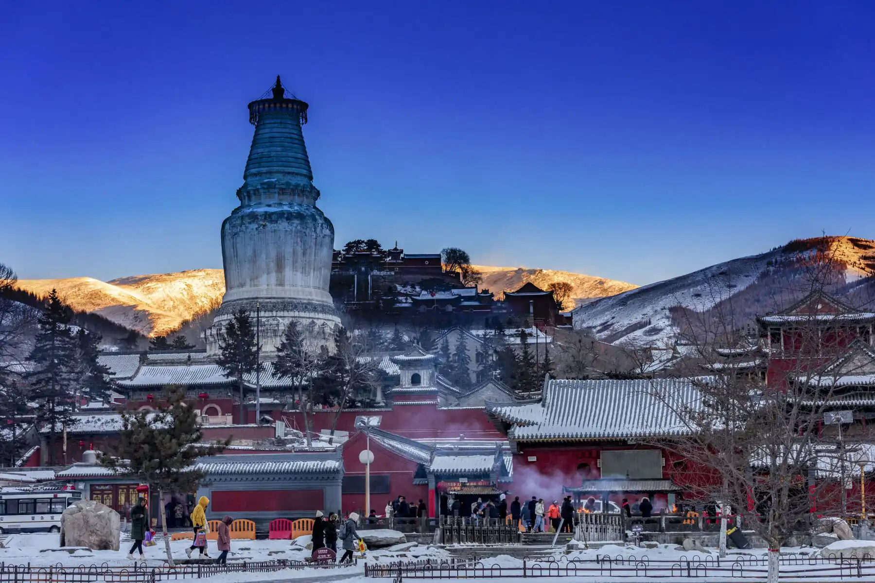 Snow-dusted White Pagoda under a Golden Mountain Sunrise, Mount Wutai, shanxi, china, china tour, Snow-covered temple complex with a towering white stupa in the foreground and snow-dusted rooftops, set against sunlit hills at dusk.