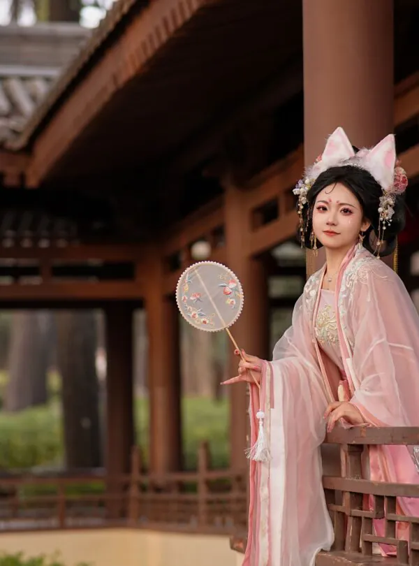 hanfu photoshoot, xi'an, female, solo, china tour, Woman in pale pink traditional attire with floral headpiece standing on a wooden veranda, holding a round embroidered fan.