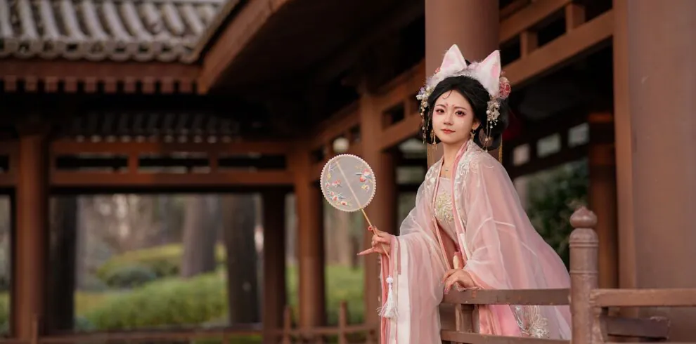 hanfu photoshoot, xi'an, female, solo, china tour, Woman in pale pink traditional attire with floral headpiece standing on a wooden veranda, holding a round embroidered fan.