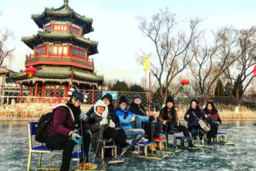 beijing pagoda scenic area, chinaexplorertour, Group of nine people in winter gear posing and smiling while seated on chairs on a frozen pond, with a traditional Chinese pagoda and red lanterns in the background.