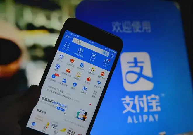 wudang retreat, taoism, hubei, china, chinaexplorertour, Close-up of a smartphone screen showing app icons, held in front of a blue Alipay sign with 支付宝, indicating mobile payments.