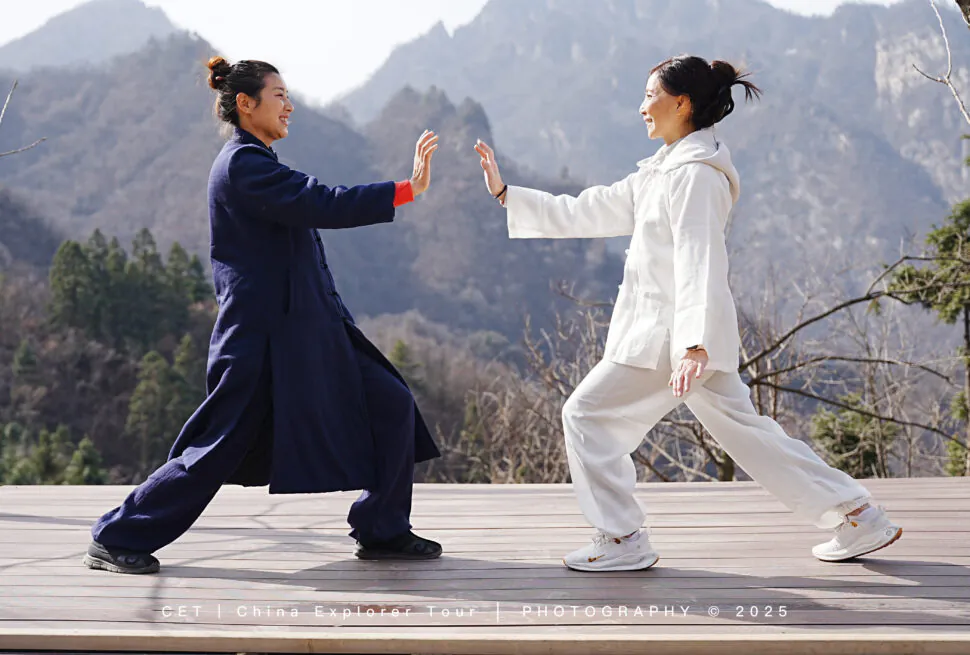 Wudang Tai Chi Retreat, hubei, china, chinaexplorertour, Two people in navy and white martial arts outfits on a wooden platform, mid high-five with a mountain landscape behind them.