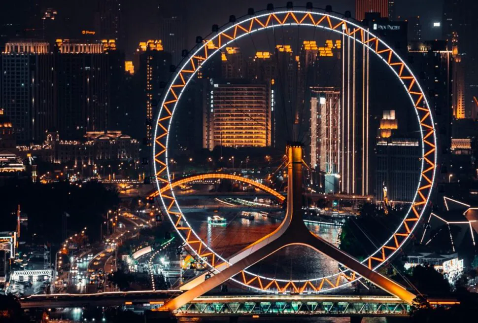 tianjing, china, chinaexplorertour, Night view of a large illuminated Ferris wheel beside a river with a glowing city skyline in the background at night.