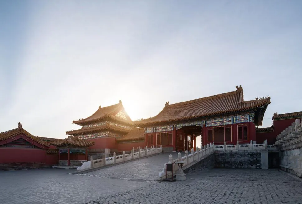 the forbidden city, beijing, china, chinaexplorertour, Ancient Chinese palace complex with red walls, ornate tiled roofs, and a stone-paved courtyard at dawn/sun behind the buildings.