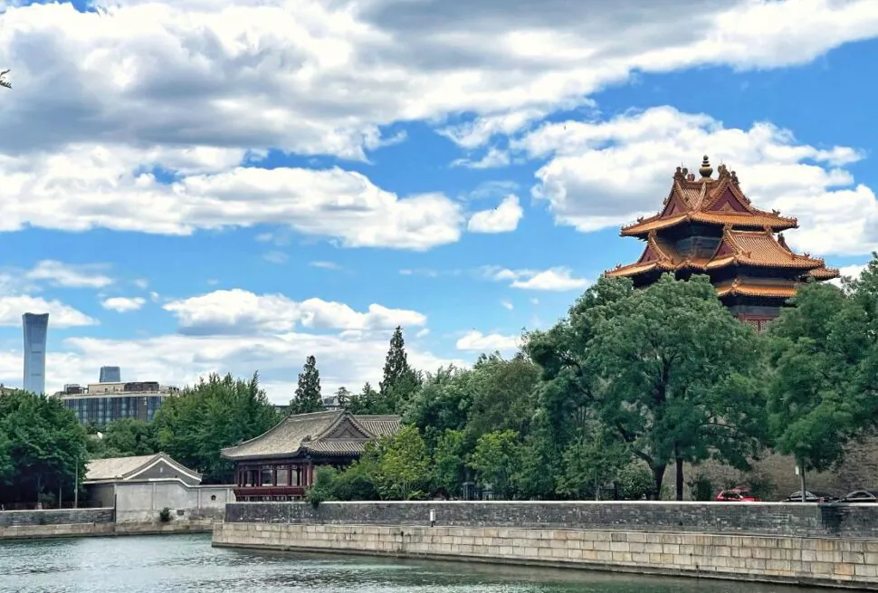 beijing, china, chinaexplorertour, Red multi-tiered pagoda surrounded by trees along a stone wall beside a river, with a modern city skyline in the distance and a blue sky above.