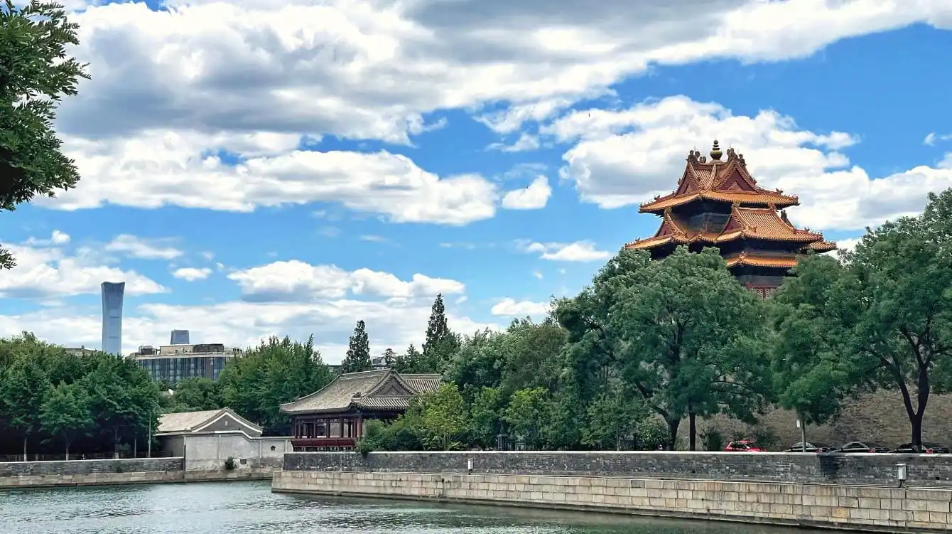 beijing, china, chinaexplorertour, Red multi-tiered pagoda surrounded by trees along a stone wall beside a river, with a modern city skyline in the distance and a blue sky above.