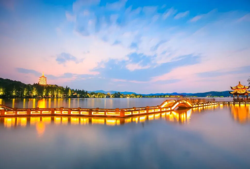 West Lake, hangzhou, zhejiang, china tour, Long-exposure view of a lit wooden boardwalk across a calm lake at twilight, with a pagoda on the left and a pavilion on the right.