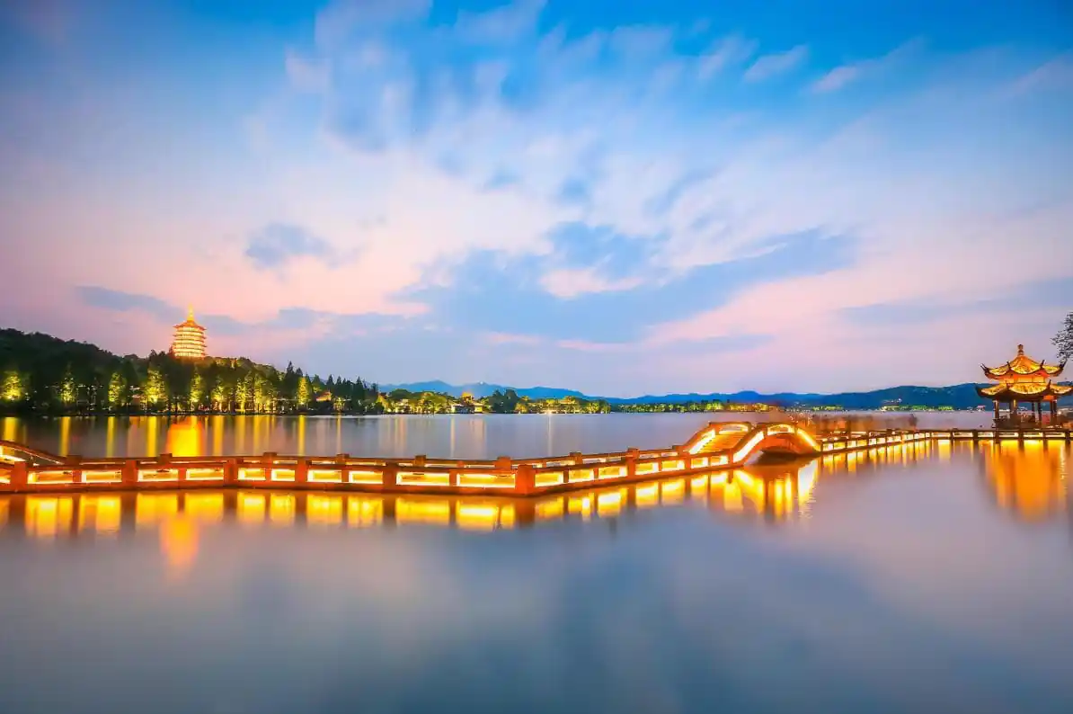 West Lake, hangzhou, zhejiang, china tour, Long-exposure view of a lit wooden boardwalk across a calm lake at twilight, with a pagoda on the left and a pavilion on the right.