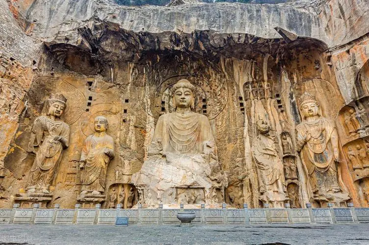 Longmen Grottoes, luoyang, henan, china tour, Rock-cut Buddhist grotto wall featuring a large central seated Buddha flanked by smaller statues, with a stone platform and railing in front.