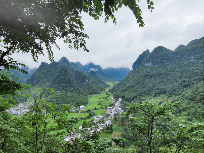 guizhou ,chinatravel, Lush green valley with a small village surrounded by steep forested mountains and a cloudy sky above.