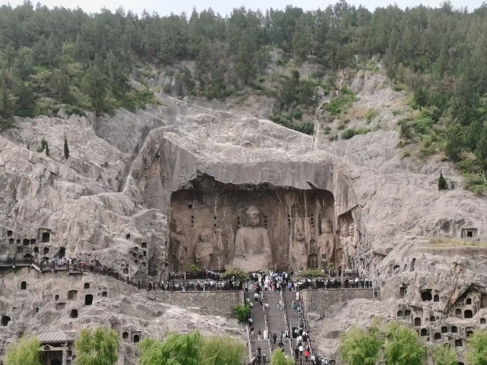 Longmen Grottoes, luoyang, henan, china tour, Rock-cut Buddhist grotto wall featuring a large central seated Buddha flanked by smaller statues, with a stone platform and railing in front., Large rock-cut Buddha statues carved into a cliff face, with a stepped walkway and crowd of visitors at the base.