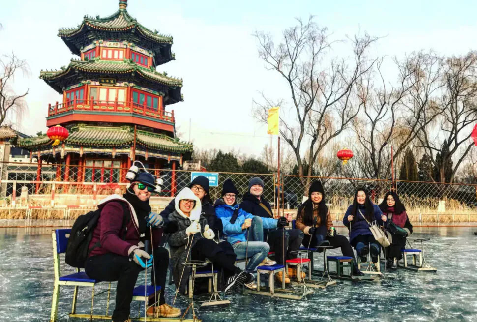 SHICHAHAI, BEIJING, CHINA TRAVEL, Group of friends in winter clothing sitting on chairs on a frozen pond, posing for a photo with a traditional Chinese pagoda and red lanterns in the background.