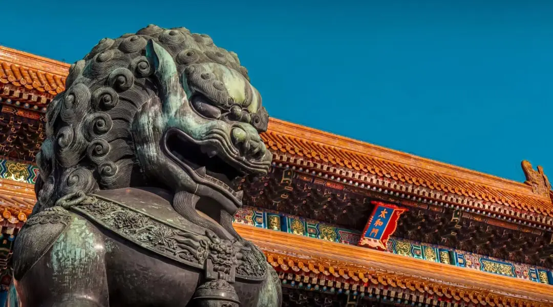 Forbidden City, beijing, china tour, Close-up of a dark stone guardian lion statue in front of a traditional Chinese temple with orange-tiled roof and colorful ornate beams against a blue sky.