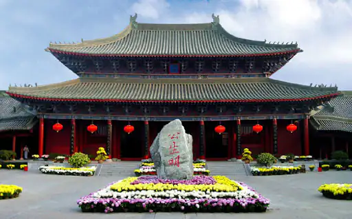 the prefecture of kaifeng, henan, chinatrip, Traditional Chinese temple with red lanterns and a central stone stele inscribed in Chinese characters, surrounded by flower beds.