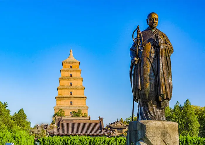 Big wild goose Pagoda, xi'an, shaanxi, chine tour, Bronze statue of a Buddhist monk holding a staff, beside a tall multi-story pagoda and traditional temple buildings in a garden under a blue sky.