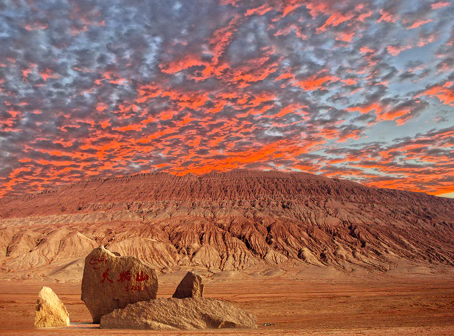 flaming mountain, turpan, xinjiang, chinatrip, Desert landscape at sunset with orange-red sky and eroded hills; a large inscribed rock and smaller boulders in the foreground.
