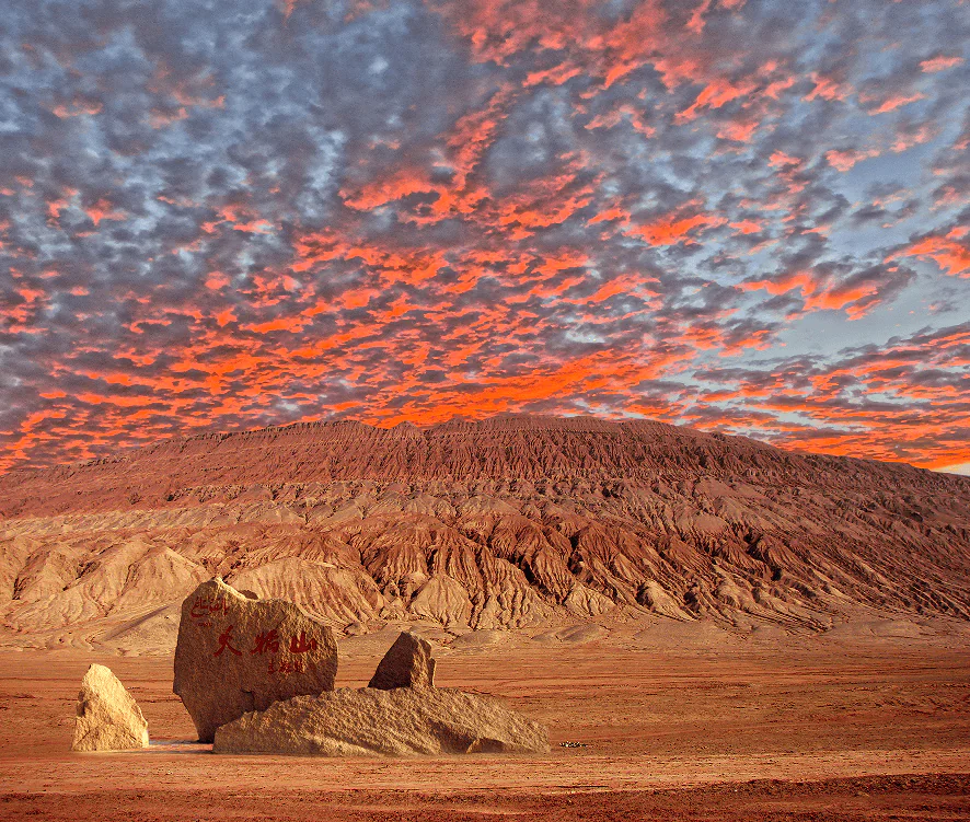 flaming mountain, turpan, xinjiang, chinatrip, Desert landscape at sunset with orange-red sky and eroded hills; a large inscribed rock and smaller boulders in the foreground.