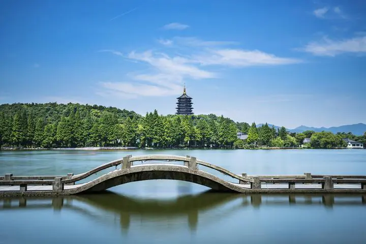 West Lake, hangzhou, zhejiang, china tour, Curved stone bridge over a calm lake with a distant multi-tiered pagoda and green trees under a blue sky.